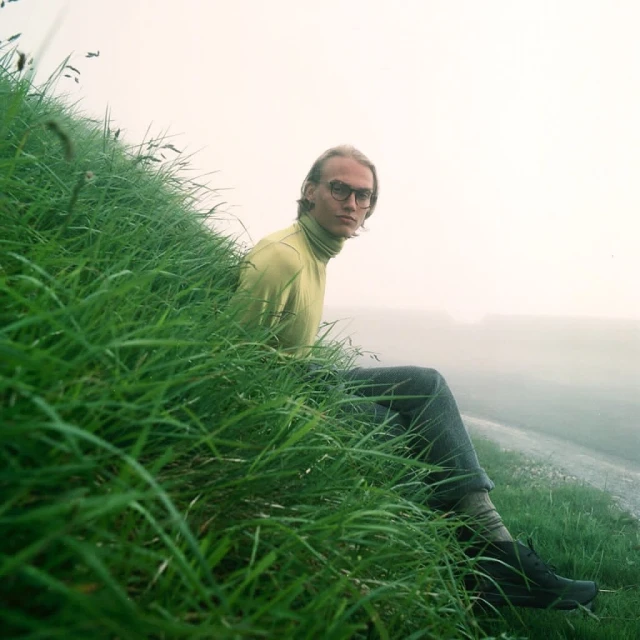 Man with glasses sits on a grassy hill in a yellow turtleneck and gray jeans, looking at the camera with fog in the distance.