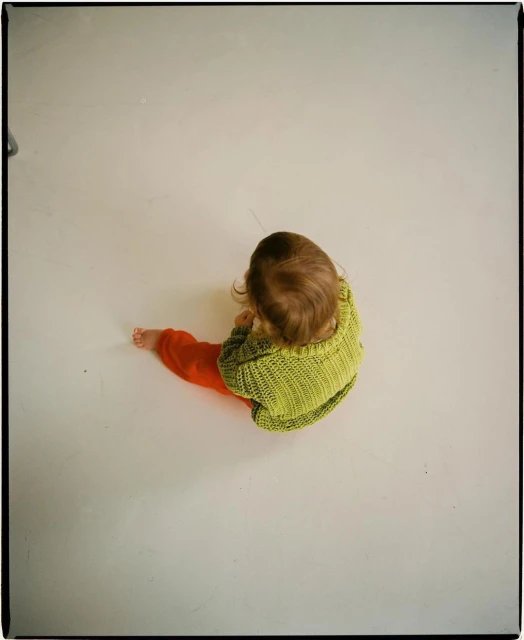 High-angle studio shot of a baby wearing a green knit sweater and orange pants, sitting on a bright white surface.