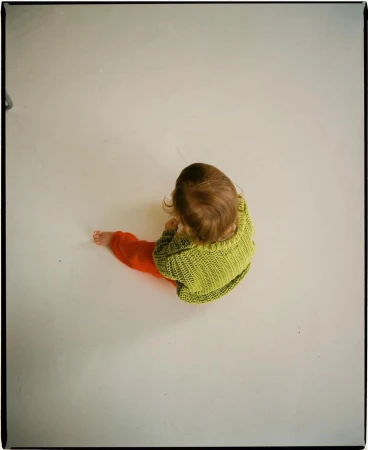 High-angle studio shot of a baby wearing a green knit sweater and orange pants, sitting on a bright white surface.