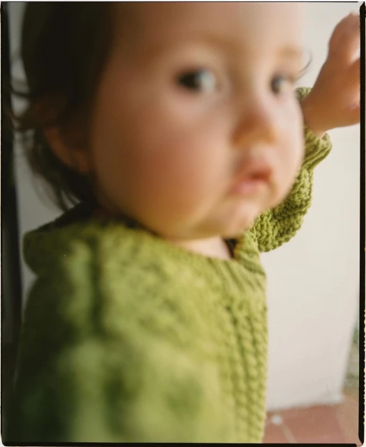 Close-up portrait of a baby with light skin and brown hair, wearing a green knitted sweater, looking to the left, with a neutral expression.
