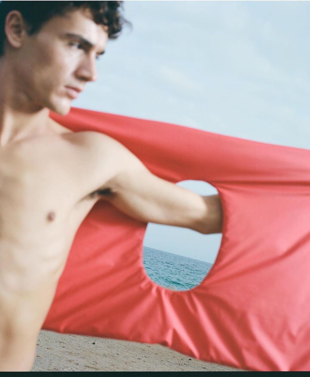 Nude man holding up a bright red fabric with a hole revealing the ocean in an outdoor setting on a sandy beach, blue sky.