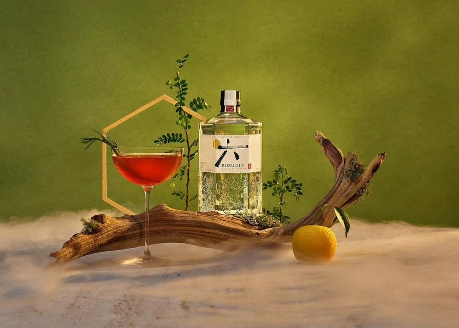 Roku Gin bottle beside an orange and red cocktail in a studio setup with green background, geometric frame, plants, and artificial fog.