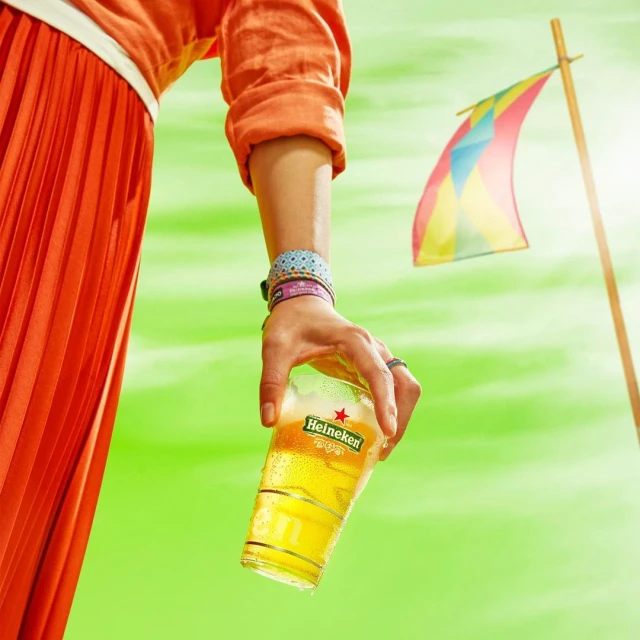 Woman in orange skirt holding an iced glass of Heineken beer against a vibrant green backdrop with a colorful flag.