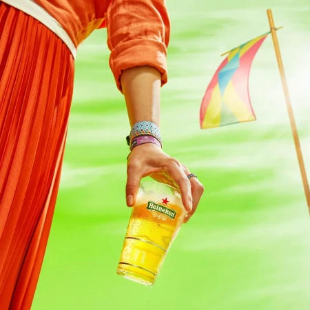 Woman in orange skirt holding an iced glass of Heineken beer against a vibrant green backdrop with a colorful flag.