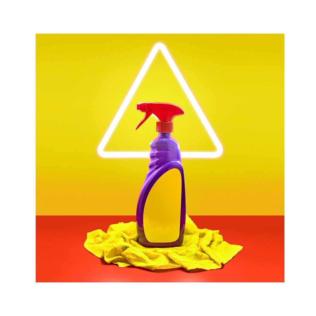Image of a purple spray bottle with a red nozzle on a yellow cloth against a yellow backdrop with a white neon triangle light.