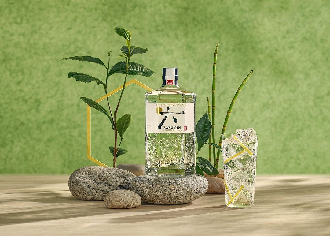 Roku Gin bottle with ice filled drink, plant sprigs, and river stones, product shot with a green backdrop and natural light.