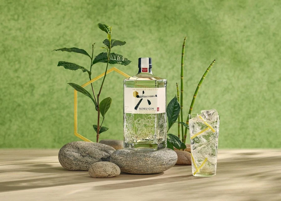 Roku Gin bottle with ice filled drink, plant sprigs, and river stones, product shot with a green backdrop and natural light.