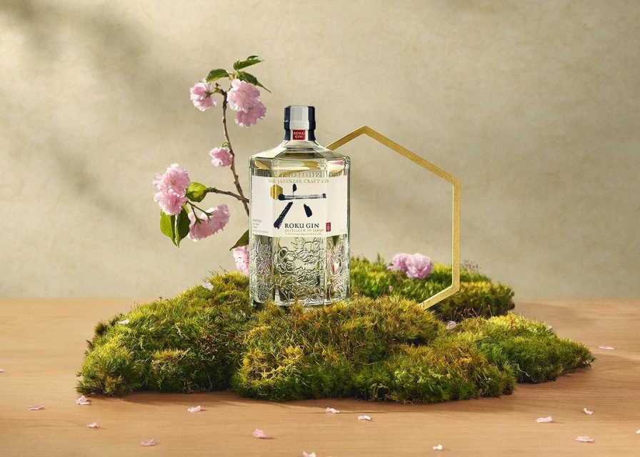 Roku Gin bottle displayed on moss with blossoming cherry branches and a gold geometric accent against a soft neutral backdrop.
