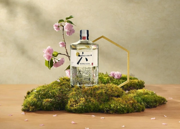 Roku Gin bottle displayed on moss with blossoming cherry branches and a gold geometric accent against a soft neutral backdrop.