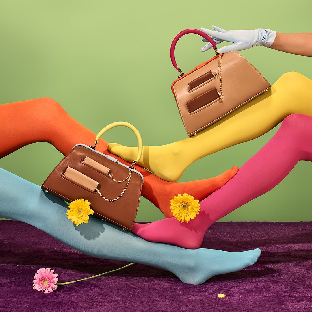 Fashion still life featuring handbags, legs in bright stockings, and flowers against a green backdrop, creating a vibrant and playful composition.