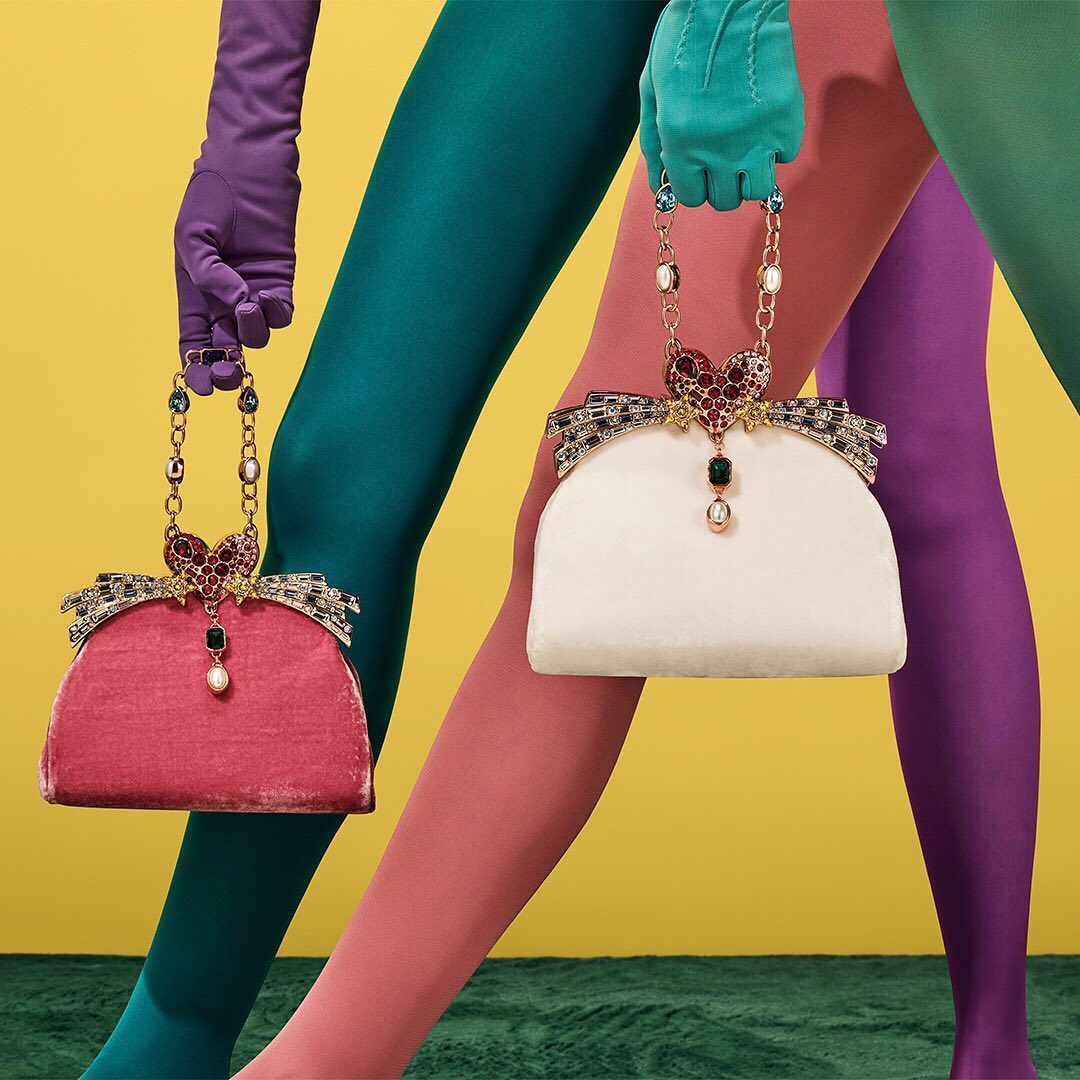 Fashion still life of two jeweled handbags held by models wearing gloves and tights in purple, teal, and pink against yellow backdrop.