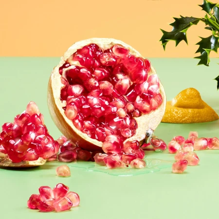 Still life shot of a halved pomegranate with loose seeds scattered around, against a yellow and green background with a lemon prop.