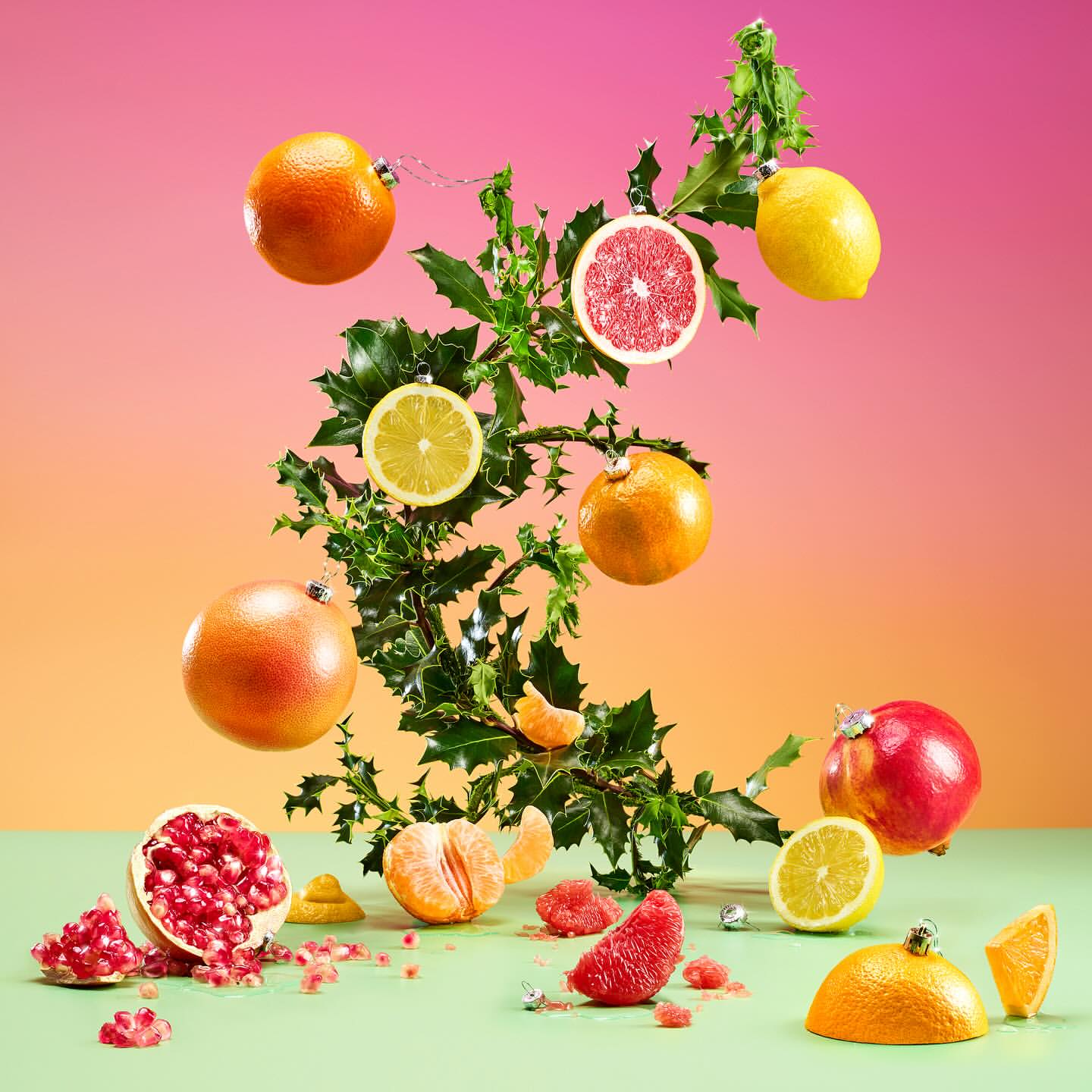 Creative still life with citrus fruit cut in half, ornamented with Christmas ball attachments, placed on a green surface against a colorful background.