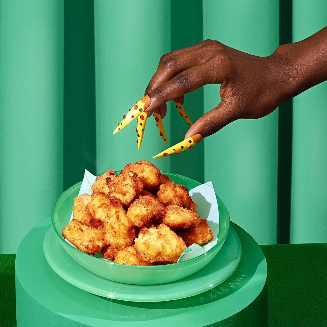 Fried food appetizers served in a green bowl held by an arm with yellow and red polka dot nails against a teal background.