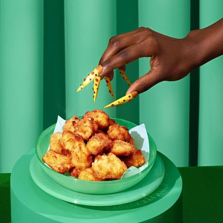 Fried food appetizers served in a green bowl held by an arm with yellow and red polka dot nails against a teal background.