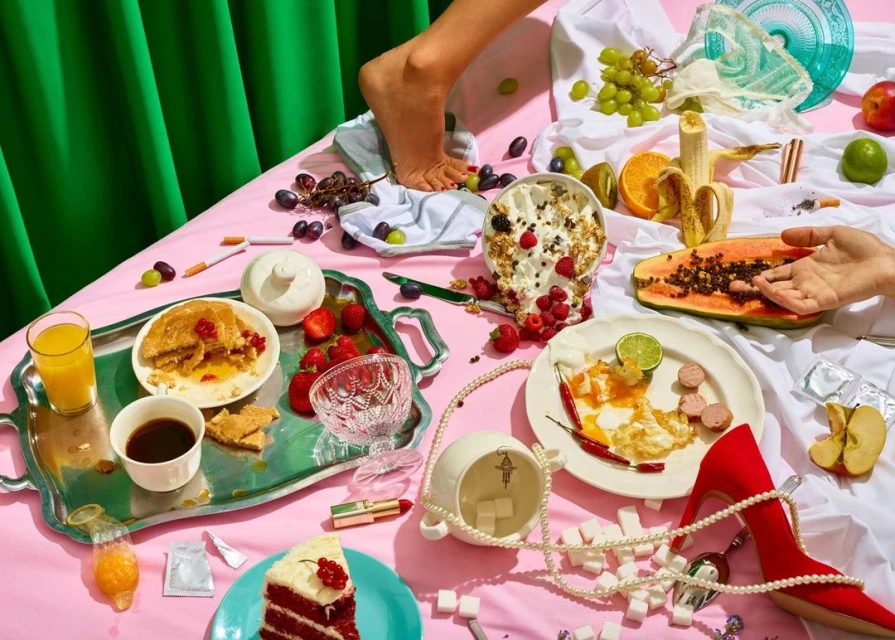 Still life featuring various foods, fruits, a red high-heeled shoe, sugar cubes, and cosmetics arranged on a pink surface with a green backdrop.