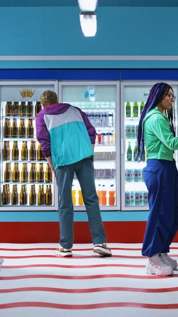 Two individuals in front of refrigerators filled with drinks, standing on a red and white striped floor, with a blue wall background.