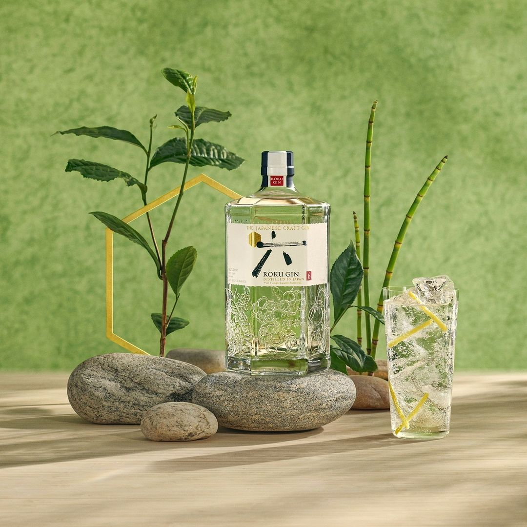 Roku Gin bottle and a glass of gin and tonic on a table with natural elements against a green backdrop, advertising.