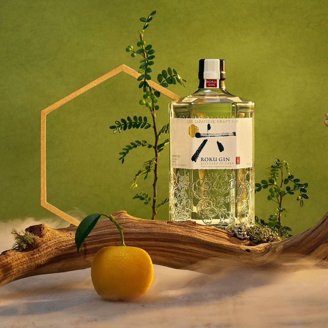 Still life shot of a clear Roku gin bottle on a wooden log with greenery, set against a green backdrop with geometric shape.