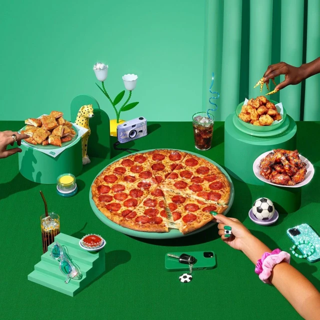 Still life featuring a pepperoni pizza, chicken bites, and various objects on a green background, with hands reaching for the food.
