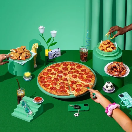 Still life featuring a pepperoni pizza, chicken bites, and various objects on a green background, with hands reaching for the food.