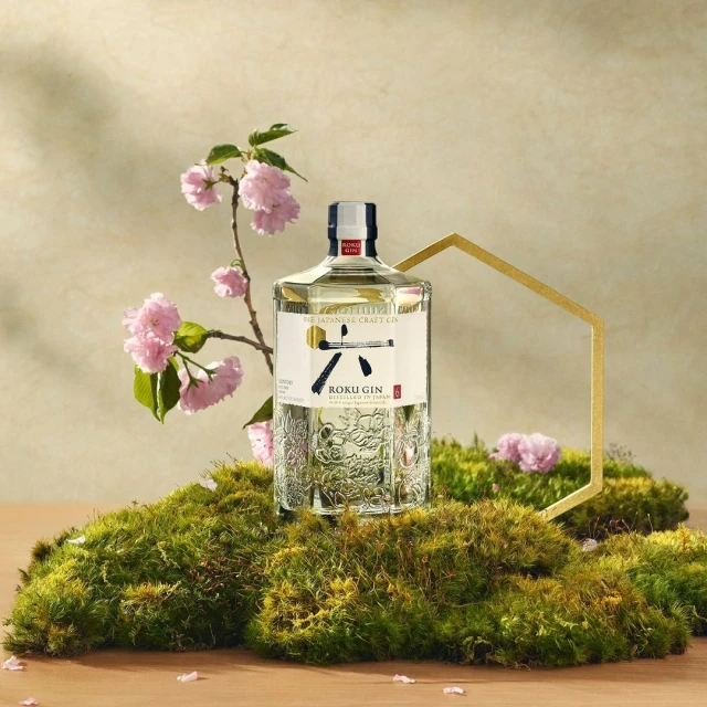Koku Gin bottle displayed on a mossy surface with pink blossoms, set against a neutral backdrop. Clean product shot for marketing.