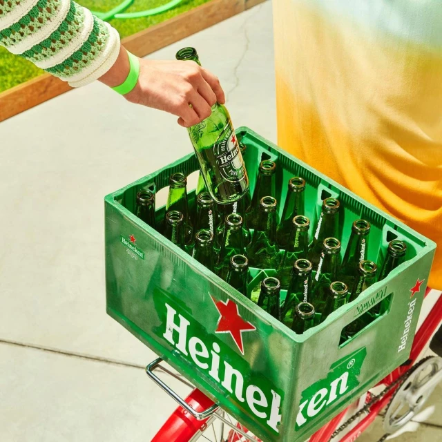 Person puts a Heineken beer bottle into a green crate filled with beer bottles, the crate is on a red bicycle outdoors.