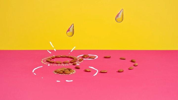 Almonds are arranged on a pink surface with white drawings, with two liquid drops hovering in front of a yellow background.  