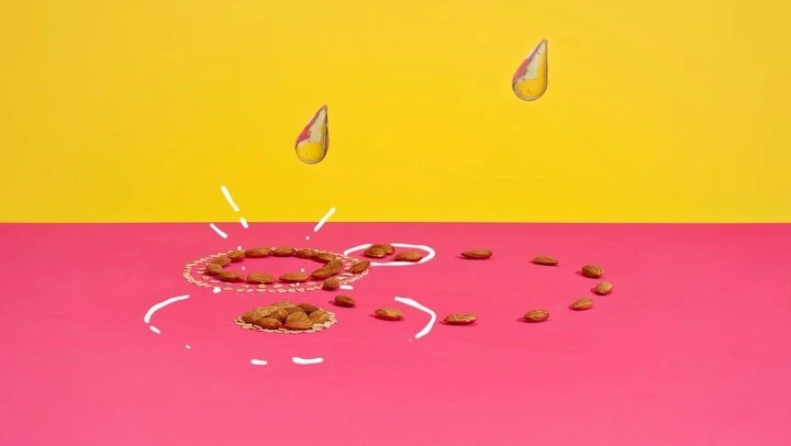 Almonds are arranged on a pink surface with white drawings, with two liquid drops hovering in front of a yellow background.  