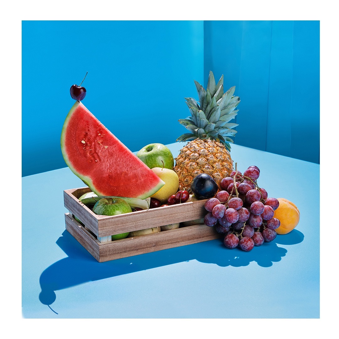 Still life shot of a wooden crate overflowing with fresh fruits, including watermelon, pineapple, grapes, and apples, against a blue backdrop.