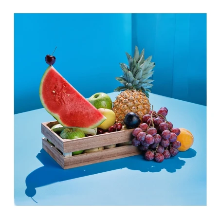 Still life shot of a wooden crate overflowing with fresh fruits, including watermelon, pineapple, grapes, and apples, against a blue backdrop.