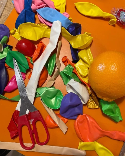 Still life with colorful uninflated balloons, metal scissors with red handles, and a bright orange on an orange surface.