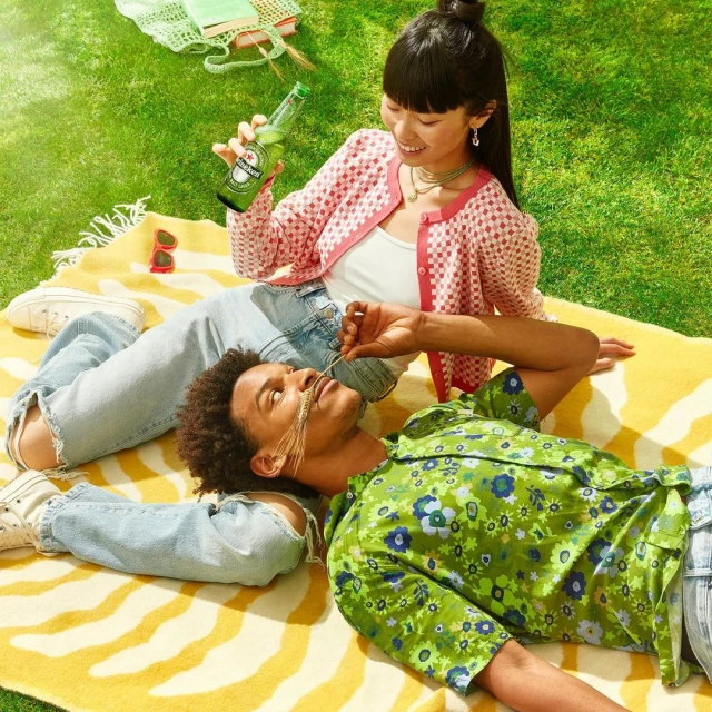 A young adult couple enjoys a sunlit picnic in the grass, complete with a patterned blanket, bottle of Heineken, and light, summery clothing.