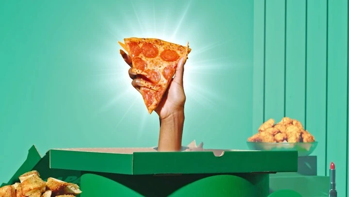 Hand holding a slice of pepperoni pizza above a pizza box with piles of garlic knots and fried chicken, set against a teal backdrop.