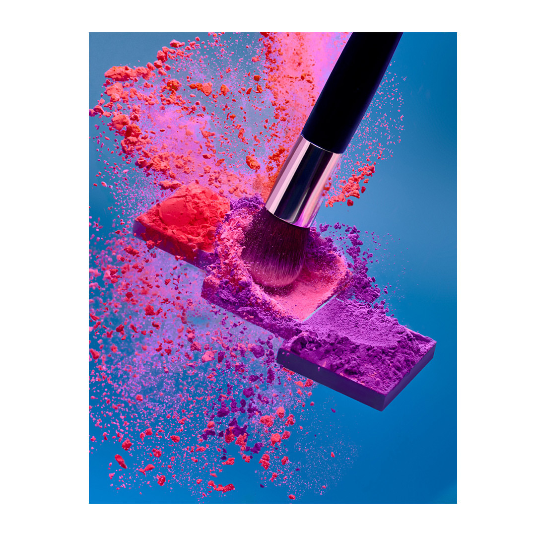 Close-up of a makeup brush touching a vibrant palette with floating, colorful powder against a striking blue background.