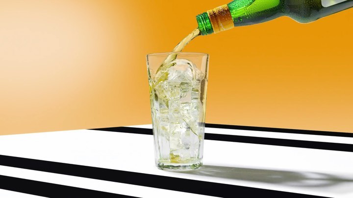 Beer being poured into tall glass filled with ice cubes, set against a white surface and a vibrant yellow gradient backdrop.