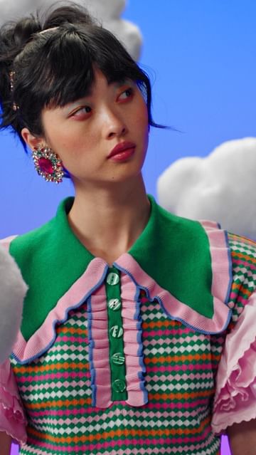 A brunette woman poses in a brightly colored green, pink, and white patterned top against a blue and white cloud-filled background.