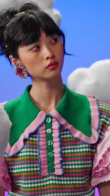 A brunette woman poses in a brightly colored green, pink, and white patterned top against a blue and white cloud-filled background.