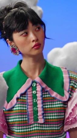 A brunette woman poses in a brightly colored green, pink, and white patterned top against a blue and white cloud-filled background.