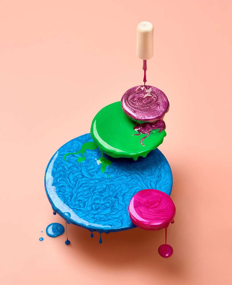 Stacked circles of nail polish in blue, green, and pink hues, with a nail polish brush hovering over the purple layer, against a peach backdrop.