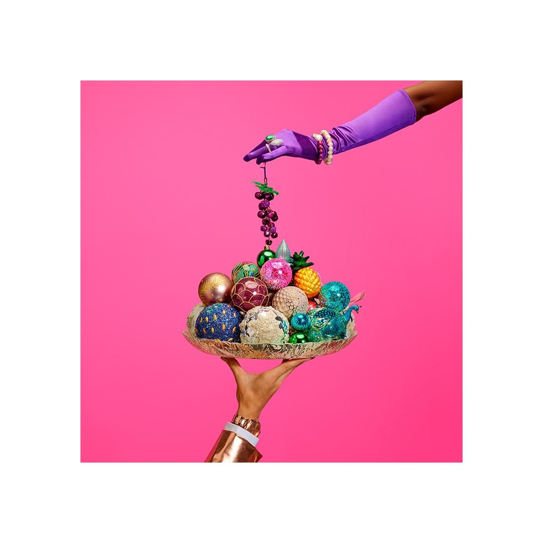 Studio shot featuring hands holding a gold tray with decorative baubles, while another hand in a purple glove holds grapes against a pink backdrop.