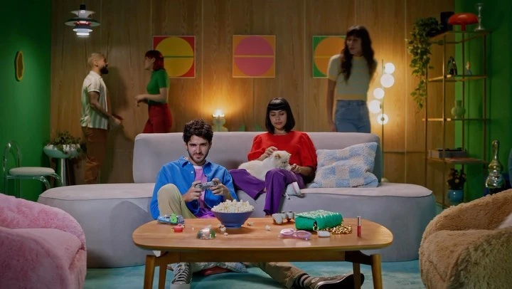 Two people are on the couch playing video games with their cat, while two other people talk in the background in a retro-styled living room.
