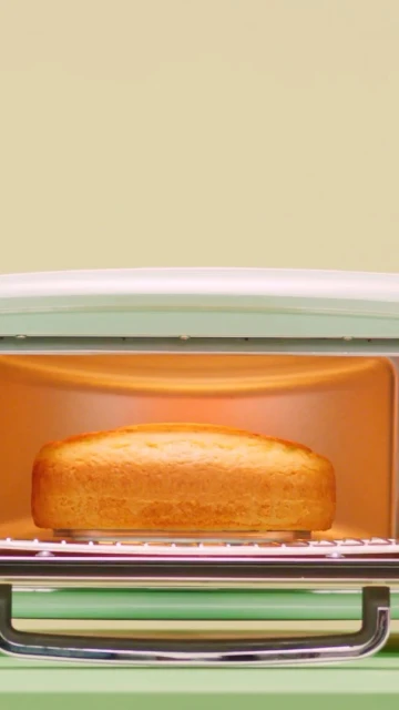 A loaf of golden-brown bread bakes inside a small, retro-style, light-green toaster oven against a light-yellow background.