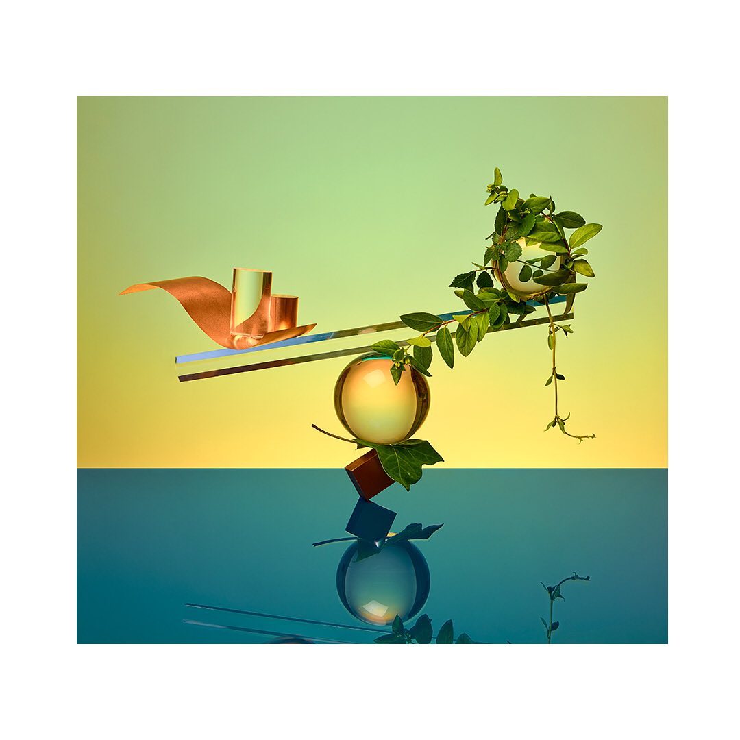 Abstract still life with floating geometric shapes, green vines, and glass spheres against a split yellow and blue background. Modern, minimal.