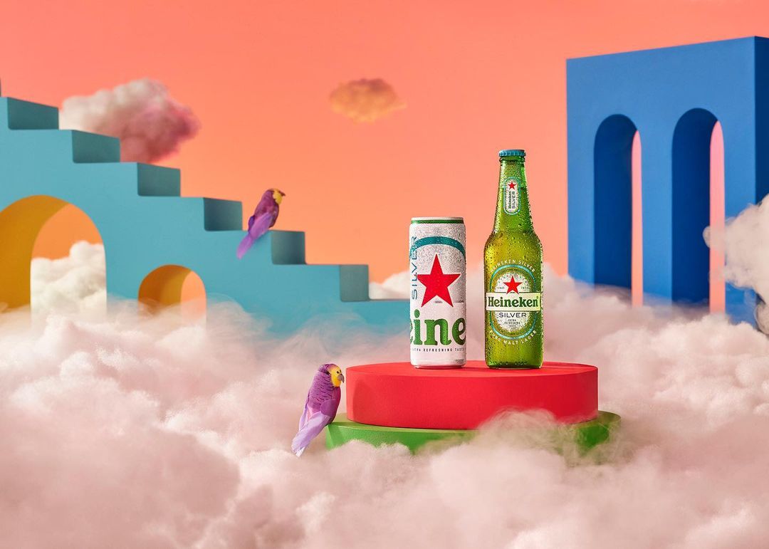 Heineken Silver products displayed on a red platform, surrounded by white clouds, birds, and a blue architectural structure.