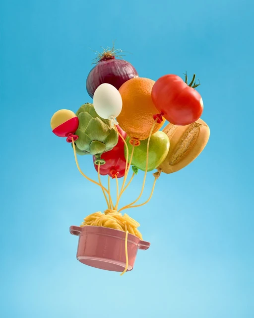 Conceptual food still life: pasta overflowing from a pot, attached to balloons of mixed vegetables, a tomato, and bread, on a blue background.
