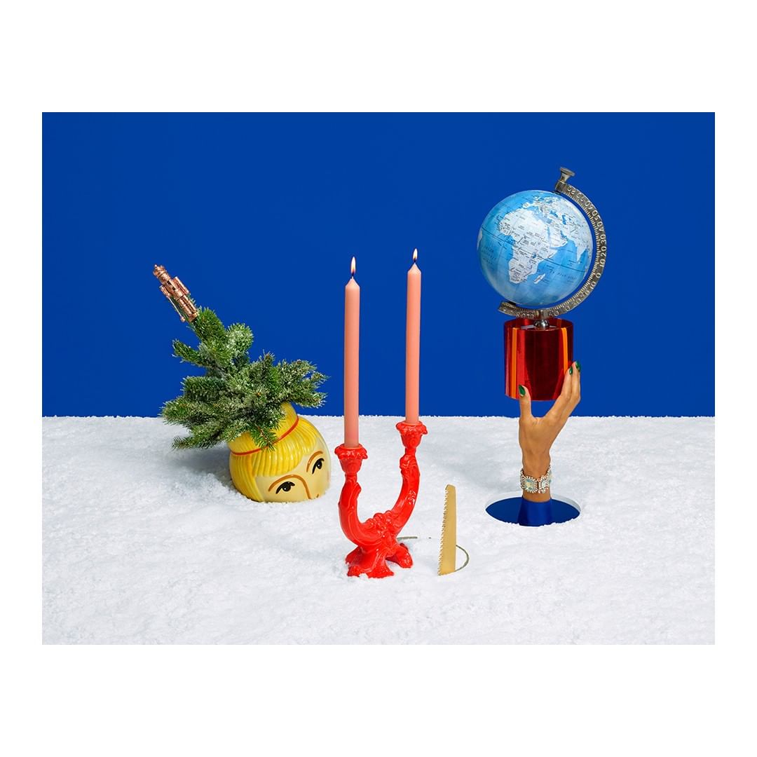 Whimsical holiday scene featuring lit candles, a vintage head vase with greenery, a globe held by a hand prop, and snow on a blue background.