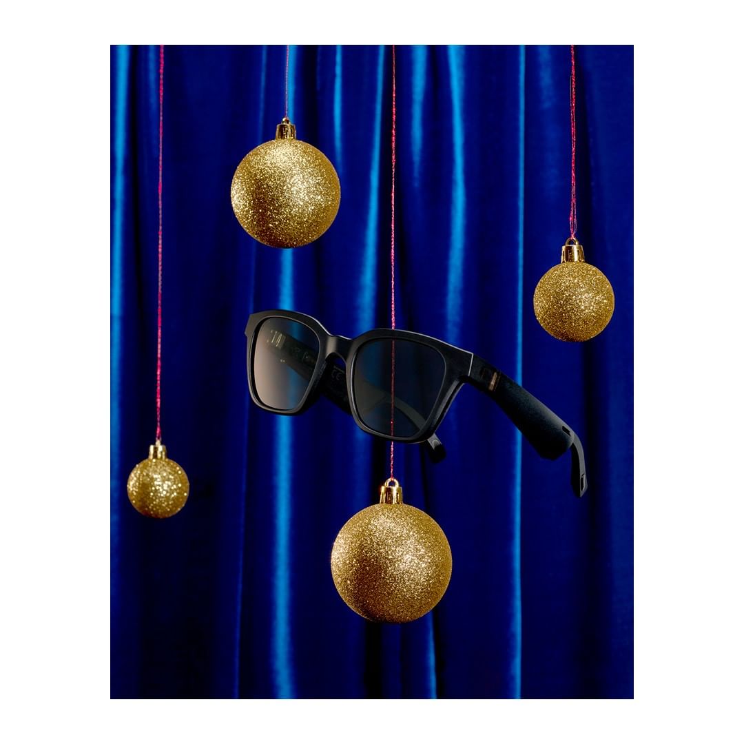 Black sunglasses float among sparkling gold Christmas ornaments against a draped royal blue fabric backdrop with dark-red dangling threads.