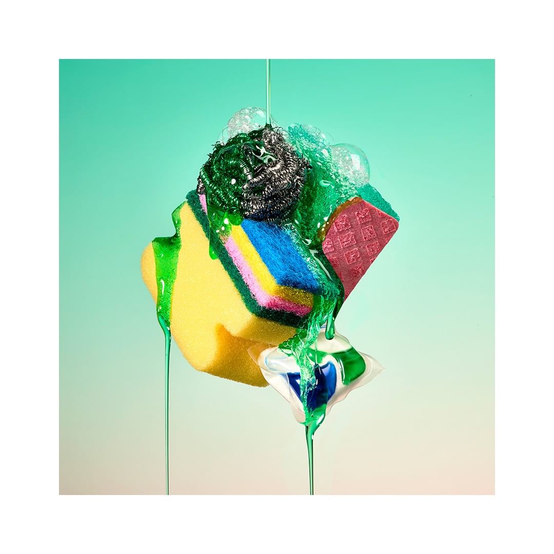 Stacked cleaning sponges and a dishwasher pod with green soap liquid flowing down against a gradient teal background in a studio shot.