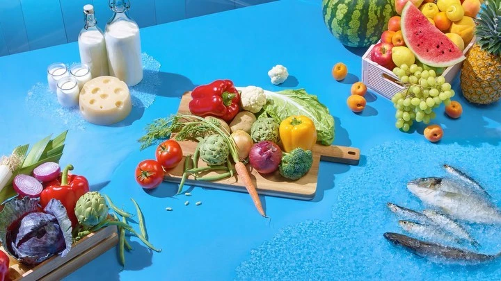 Overhead shot of fresh produce and dairy products including fish, vegetables, fruits, cheese, and milk arranged on a blue surface.